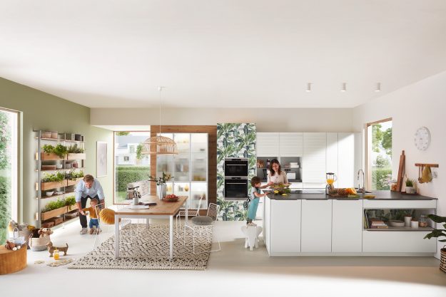 Familie in moderner Küche – Mutter kocht mit Kind auf der Kücheninsel, Vater spielt mit Kleinkind.